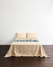 A neatly made bed with a beige quilt cover in Lagoon & Creme with matching pillowcases in Creme, set against a plain white wall and dark wood flooring. - view