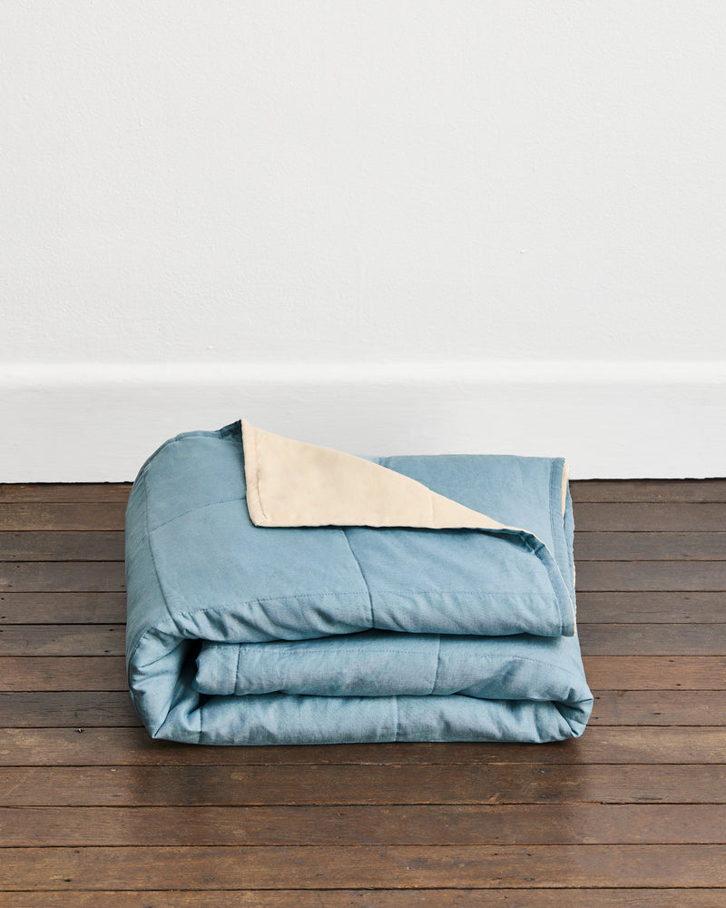 A folded dusty blue quilt cover in Lagoon & Creme with a creme underside, displayed on a dark wooden floor against a white wall.