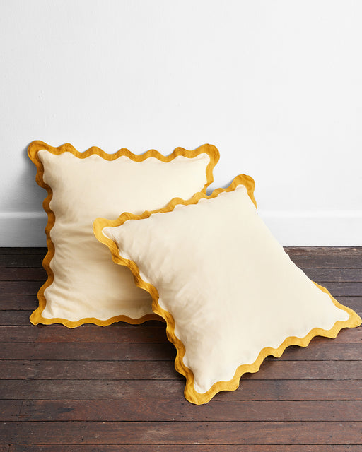 The Limoncello & Turmeric 100% French Flax Linen Scalloped European Pillowcases (Set of Two) by Bed Threads add charm to the room, their wavy yellow edges contrasting beautifully against the dark floor and white wall. One pillow leans on the other.
