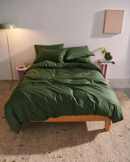 A bed topped with the Moss Organic Cotton Flat Sheet by Bed Threads features green pillows on a beige rug. Pink nightstands with books, a lamp, and a plant flank the bed. Above, rectangular artwork decorates the white wall.