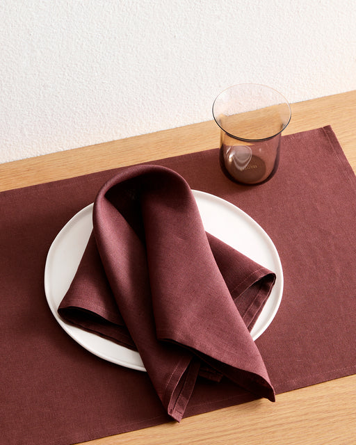 A white plate with a folded Bed Threads Cacao 100% French Flax Linen Napkin rests on a matching placemat atop a wooden table, with a transparent brown glass placed beside the plate.