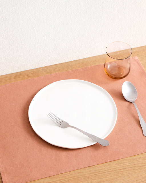 A white plate with a fork sits on a Hazelnut 100% French Flax Linen Placemat by Bed Threads, beside a spoon and an amber glass, all arranged on a light wooden table for stylish table protection.