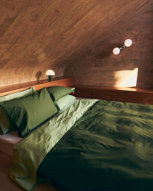 A cozy attic bedroom with slanted wooden walls, a bed dressed in Bed Threads’ Apple Organic Cotton Flat Sheet, two modern lamps—one on the headboard and one wall-mounted—and sunlight filtering through a window.