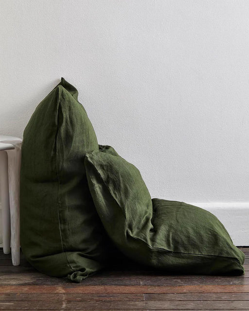 Two large, dark green linen floor cushions evoke the Olive & Oatmeal & White Stripe 100% French Flax Linen Bedding Bundle by Bed Threads, stacked on a wooden floor beside a white wall and near a partially visible white chair.