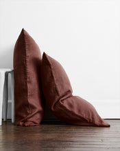 Two Cacao 100% French Flax Linen Pillowcases by Bed Threads rest against a white wall and wooden floor—one upright, one angled. A small white side table appears on the left. - view