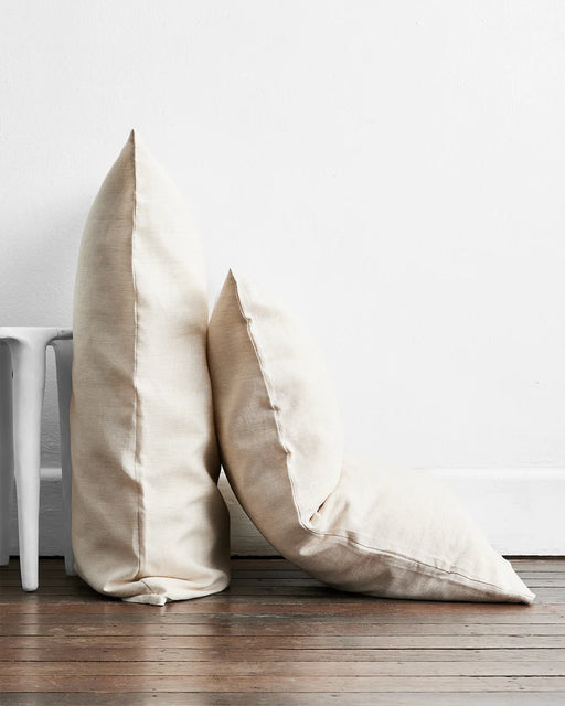 Two off-white pillows from the Bed Threads Olive & Oatmeal & White Stripe 100% French Flax Linen Bedding Bundle rest against a white wall on a wooden floor—one upright, one leaning, with part of a white stool visible on the left.