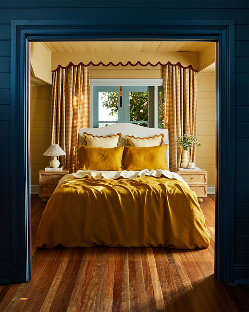 A cozy bedroom features Bed Threads' Limoncello & Turmeric 100% French Flax Linen Scalloped European Pillowcases, yellow bedding, wicker nightstands with lamps and vases, tall checked curtains above the bed, and warm wood floors.