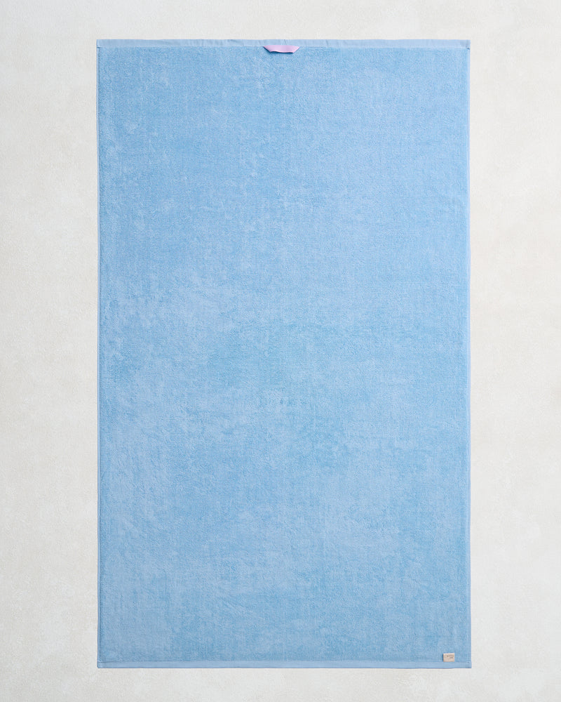 The Sky 100% Cotton Terry Bath Sheet by Bed Threads features a rectangular light blue design with a purple loop and beige tag—an ideal plush, quick-drying towel to elevate any bathroom.