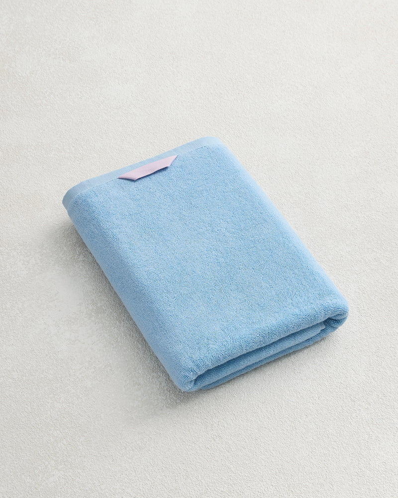 The Bed Threads Sky 100% Cotton Terry Bath Towel with a pink tag is folded on a textured white surface, highlighting its plush softness and premium quality.