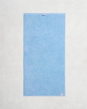 A pair of rectangular, light blue Sky 100% Cotton Terry Bath Towels by Bed Threads are laid flat on a textured surface, each featuring a small fabric loop at the top edge and a beige tag on the bottom right corner. - view