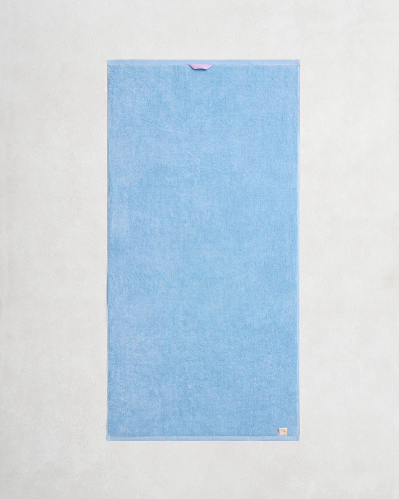 A pair of rectangular, light blue Sky 100% Cotton Terry Bath Towels by Bed Threads are laid flat on a textured surface, each featuring a small fabric loop at the top edge and a beige tag on the bottom right corner.