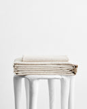 A stack of Bed Threads' Oatmeal & White Stripe 100% French Flax Linen Sheet Set rests on a white stool against a plain background, showcasing the elegance and temperature-regulating properties of this premium bedding.