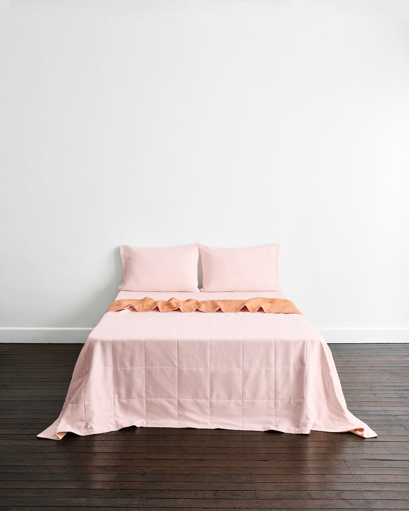 A neatly made bed with a light pink quilt cover in Terracotta & Rosewater with two matching pillowcases in Rosewater set against a plain white wall and dark wood floor.