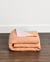 A folded peach-colored quilt cover in Terracotta & Rosewater with a light pink underside in Rosewater, displayed on a dark wood floor against a white wall. - view