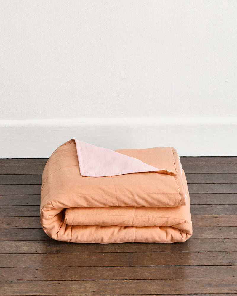 A folded peach-colored quilt cover in Terracotta & Rosewater with a light pink underside in Rosewater, displayed on a dark wood floor against a white wall.