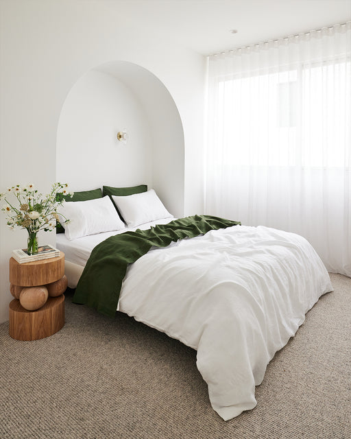 A minimalist bedroom features a bed dressed in white linens and the Olive 100% French Flax Linen Flat Sheet by Bed Threads, set under an arched alcove. Wooden side tables with flowers and sheer curtains create a serene, light-filled space.