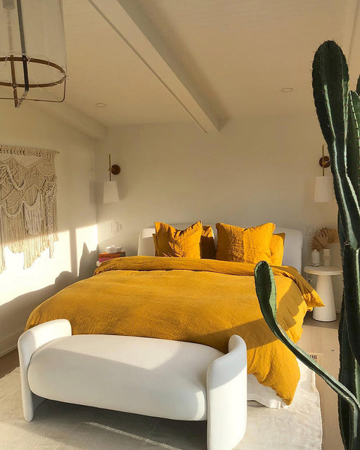 A bright, modern bedroom featuring the Bed Threads Turmeric 100% French Flax Linen Duvet Cover Set, two matching pillowcases, a white bench at the foot of the bed, macramé wall art, white walls, a tall cactus, and warm sunlight.