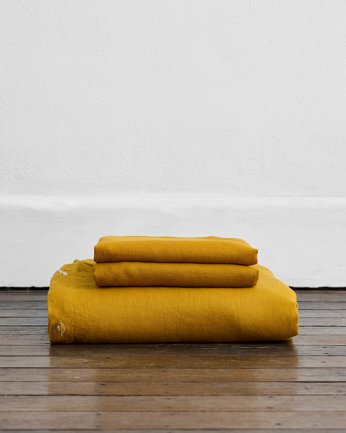 The Bed Threads Turmeric 100% French Flax Linen Duvet Cover Set, with matching pillowcases neatly folded and stacked, rests on a wooden floor against a plain white wall.