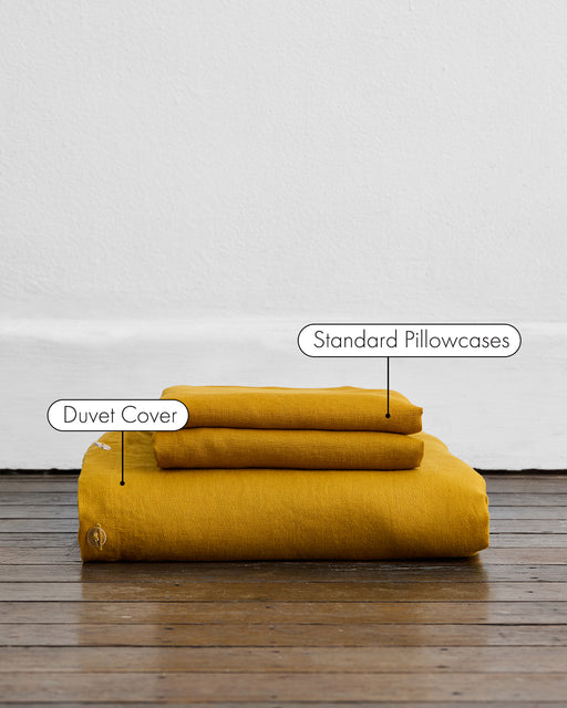 The Bed Threads Turmeric 100% French Flax Linen Duvet Cover Set, featuring a mustard yellow duvet cover and two matching pillowcases, is folded and stacked on a wooden floor against a white wall with labeled items.