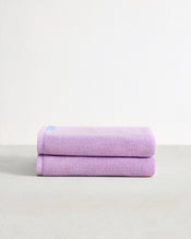 The Bed Threads Violet 100% Cotton Terry Bath Sheets (Set of Two) are stacked neatly on a light gray surface with a white background, with a small blue tag visible on the top sheet. - view