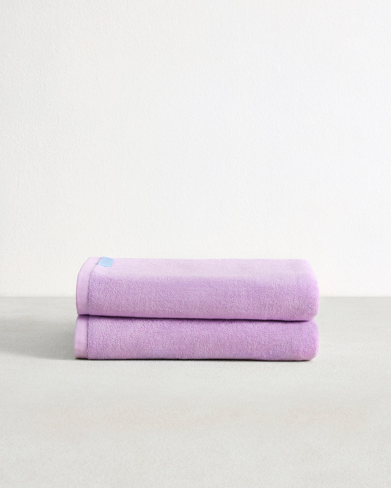 The Bed Threads Violet 100% Cotton Terry Bath Sheets (Set of Two) are stacked neatly on a light gray surface with a white background, with a small blue tag visible on the top sheet.
