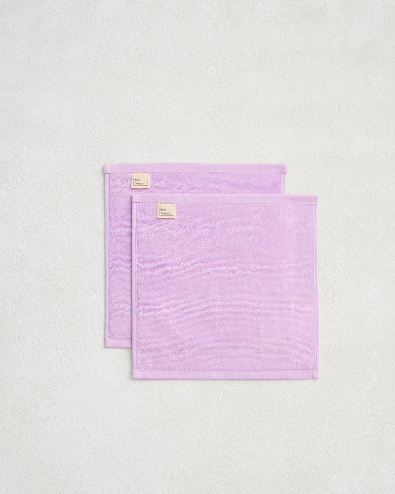 The Bed Threads Violet 100% Cotton Terry Face Washers (Set of Two) are neatly folded on a textured surface, highlighting their absorbent, skin-friendly terry fabric and subtle beige tags.