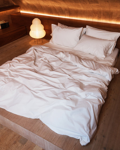 A cozy bed features the Bed Threads Crisp White Organic Cotton Sheet Set and pillows on a wooden platform in a warmly lit, wood-paneled room, complemented by a modern paper lantern lamp in the corner.