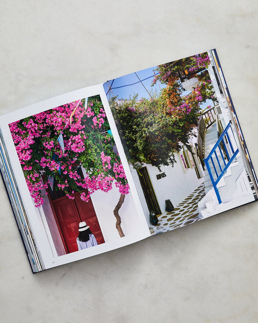 An open book on marble displays photos of a woman in a hat beneath pink bougainvillea and sunlit Cycladic streets, perfectly capturing island vibes—ideal for the Assouline Mykonos Muse by Lizy Manola.