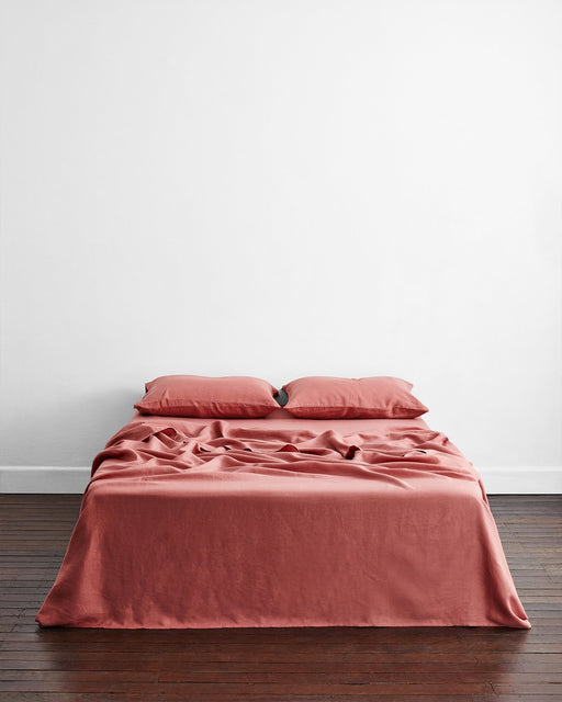 A neatly made bed with wrinkled Bed Threads Pink Clay 100% French Flax Linen Flat Sheet and two matching pillows, set against a plain white wall on a dark wooden floor.