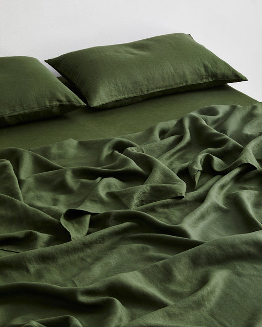 A neatly made bed with two pillows, all dressed in the Bed Threads Olive 100% French Flax Linen Flat Sheet. The slightly rumpled blanket enhances the cozy, inviting look.