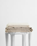 The Bed Threads Oatmeal 100% French Flax Linen Sheet Set is folded atop a sleek white stool against a plain backdrop, highlighting its temperature-regulating benefits in an elegant, minimalist display.