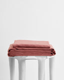 A neatly folded stack of Bed Threads Pink Clay 100% French Flax Linen Sheet Set rests on a modern white stool set against a plain white background.