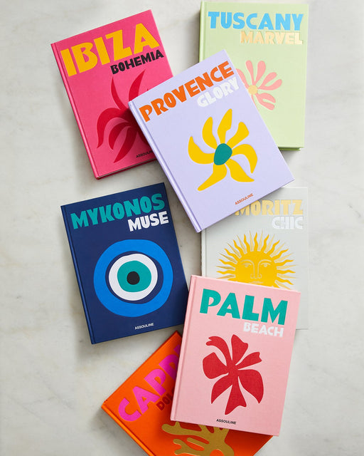 Seven vibrant Assouline books—including Mykonos Muse by Lizy Manola, Ibiza Bohemia, Provence Glory, Palm Beach, Capri Dolce Vita, St. Moritz Chic, and Tuscany Marvel—are artfully arranged on a marble surface.